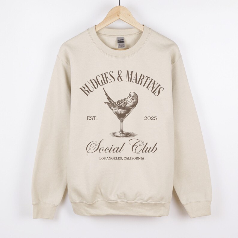 May include: A beige sweatshirt with a brown graphic print of a budgie in a martini glass. The text on the graphic reads "Budgies & Martinis Social Club Est. 2025 Los Angeles, California."