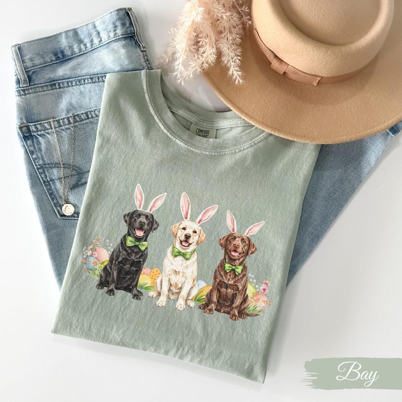 May include: A green t-shirt with a graphic of three Labrador Retrievers wearing bunny ears and bow ties. The dogs are surrounded by Easter eggs. The text "Bay" is visible in the bottom right corner of the image.