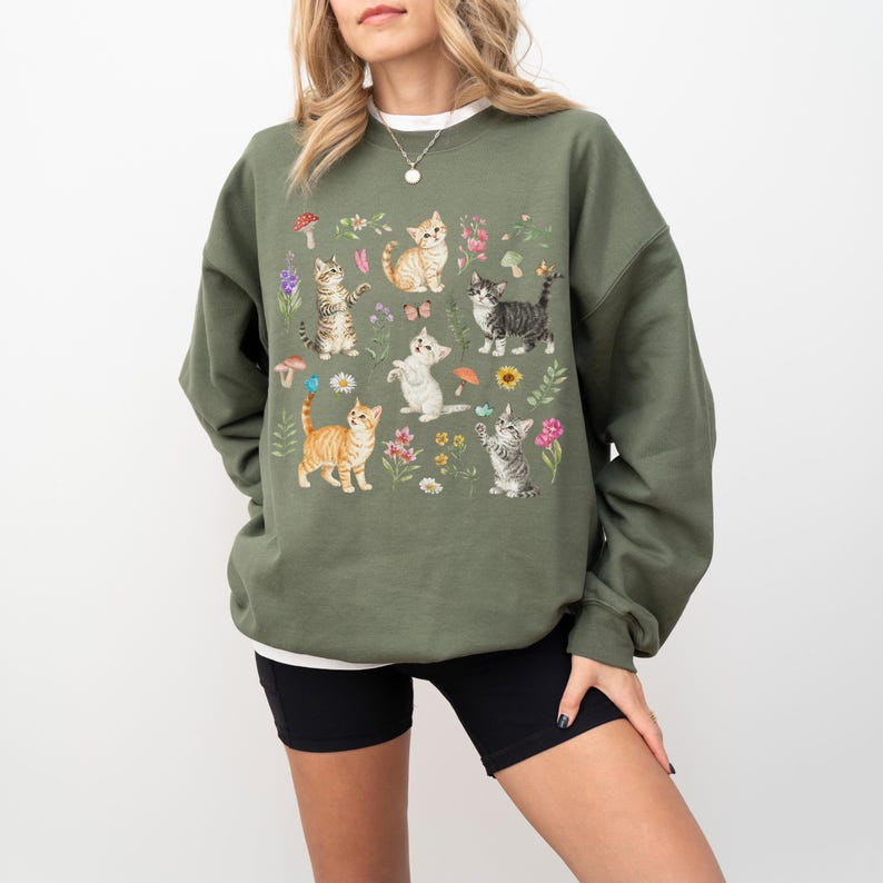 May include: A green sweatshirt with a graphic print of multiple cats and flowers. The cats are in various poses and colors, including orange, gray, and white. The flowers are in shades of pink, purple, and yellow.
