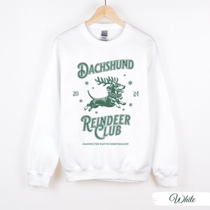 May include: White sweatshirt featuring a green graphic of a dachshund wearing a reindeer antler headband. The text "Dachshund Reindeer Club" is printed below the image. The text "Leading the way to Christmas joy" is printed below the club name.