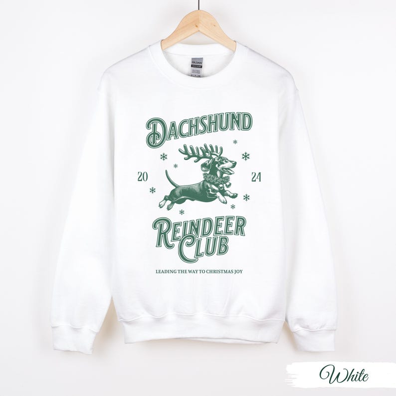 May include: White sweatshirt featuring a green graphic of a dachshund wearing a reindeer antler headband. The text "Dachshund Reindeer Club" is printed below the image. The text "Leading the way to Christmas joy" is printed below the club name.
