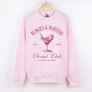 May include: Pink sweatshirt with a graphic design of a budgie in a martini glass. The text reads "Budgies & Martinis Social Club Est. 2025 Los Angeles, California."