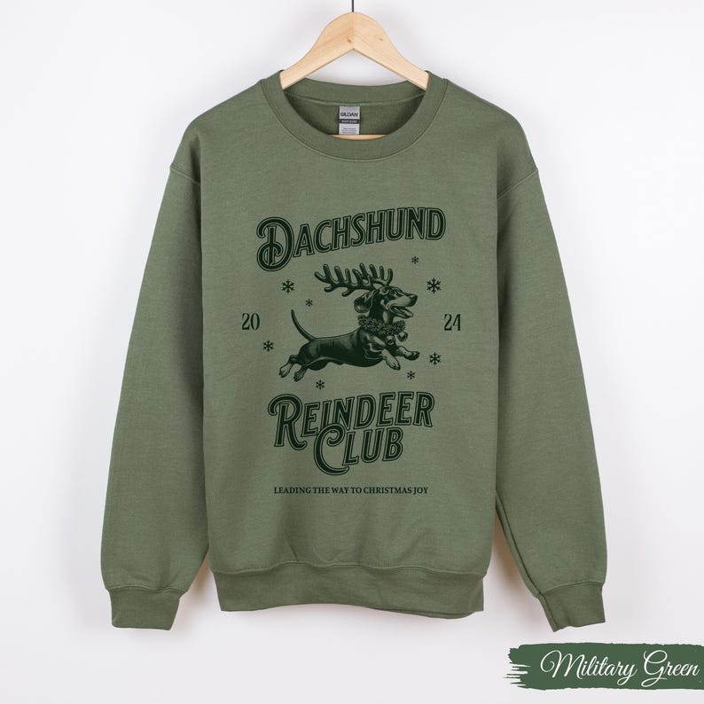 May include: A green sweatshirt featuring a graphic of a dachshund wearing a reindeer antler headband and the text "Dachshund Reindeer Club" and "Leading the way to Christmas joy".