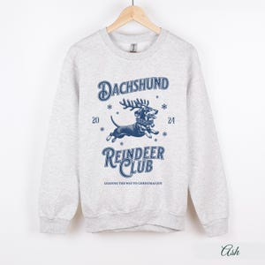 May include: A grey sweatshirt featuring a blue graphic of a dachshund wearing reindeer antlers and a Christmas wreath. The text "Dachshund Reindeer Club" is printed below the image, with the tagline "Leading the way to Christmas joy".