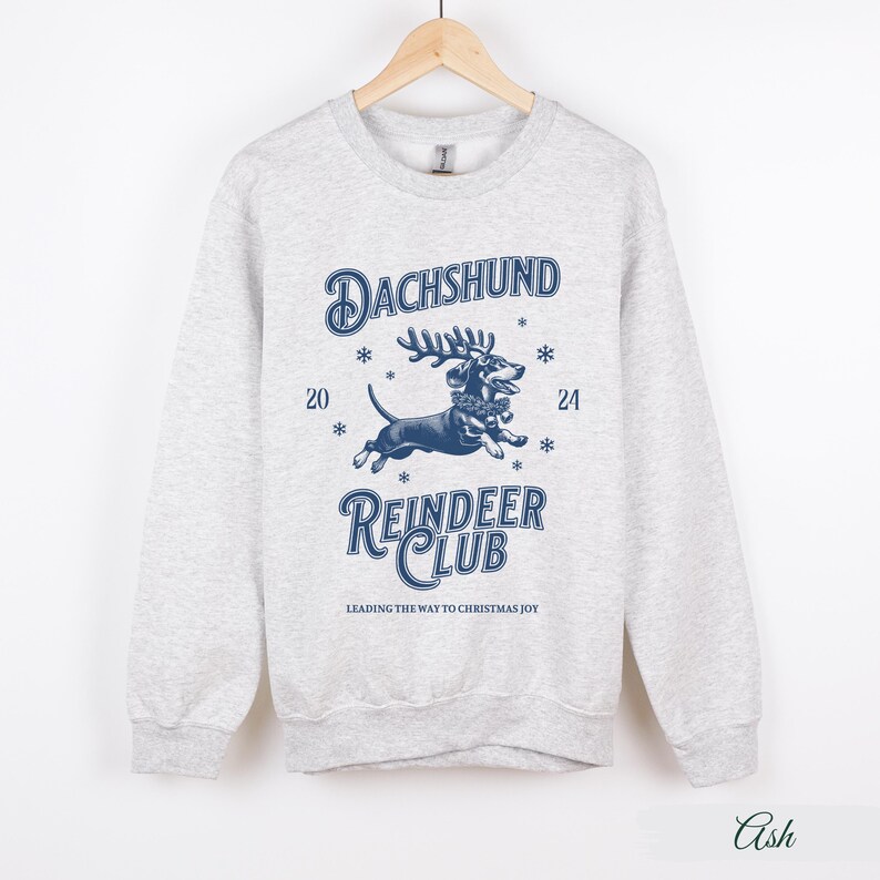 May include: A grey sweatshirt featuring a blue graphic of a dachshund wearing reindeer antlers and a Christmas wreath. The text "Dachshund Reindeer Club" is printed below the image, with the tagline "Leading the way to Christmas joy".