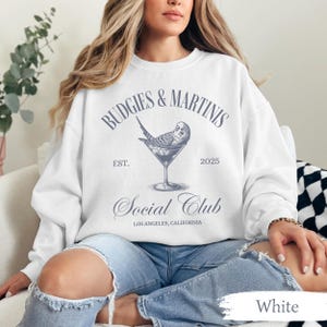 May include: White sweatshirt with a blue graphic design that says "Budgies & Martinis Est. 2025 Social Club Los Angeles, California". The graphic also features a bird in a martini glass.