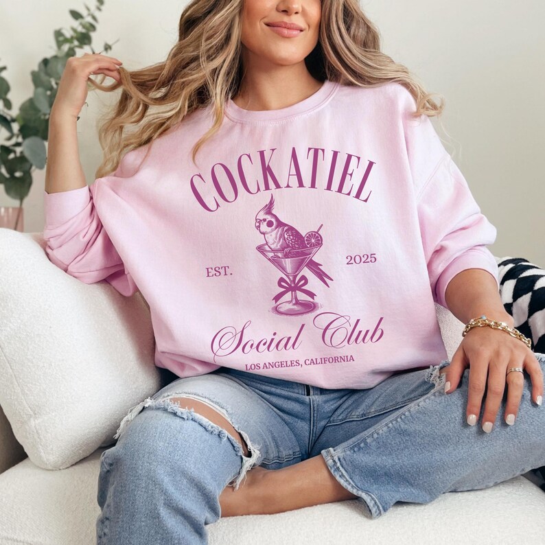May include: A pink sweatshirt with the text "Cockatiel Social Club Est. 2025 Los Angeles, California" and a pink illustration of a cockatiel in a martini glass.