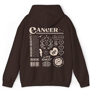 May include: Brown hooded sweatshirt with a graphic print of the zodiac sign Cancer. The print includes the words "Cancer", "Cosmic Energy", "Moon Water", "Love Yourself", and "Positive".