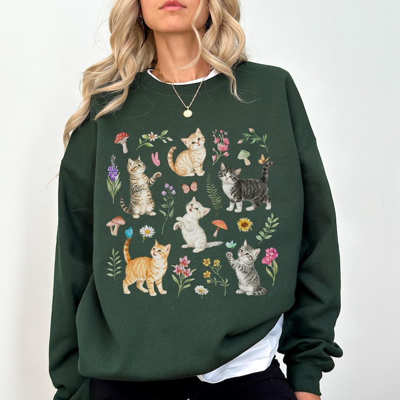May include: A dark green sweatshirt with a graphic print of multiple kittens surrounded by flowers and mushrooms. The kittens are in various poses, including sitting, standing, and playing.