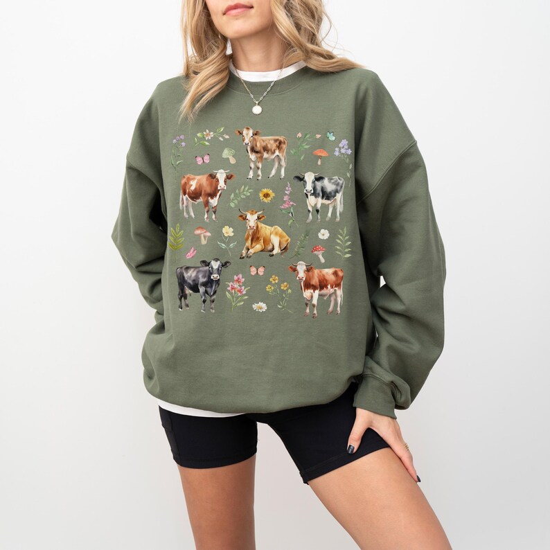 Pu&ograve; includere: Una felpa verde con una stampa di mucche, fiori e funghi. Le mucche sono in varie pose e colori.