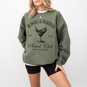May include: A green sweatshirt with the text "Budgies & Martins Social Club Est. 2025 Los Angeles, California" and a black and white illustration of a budgie in a martini glass.