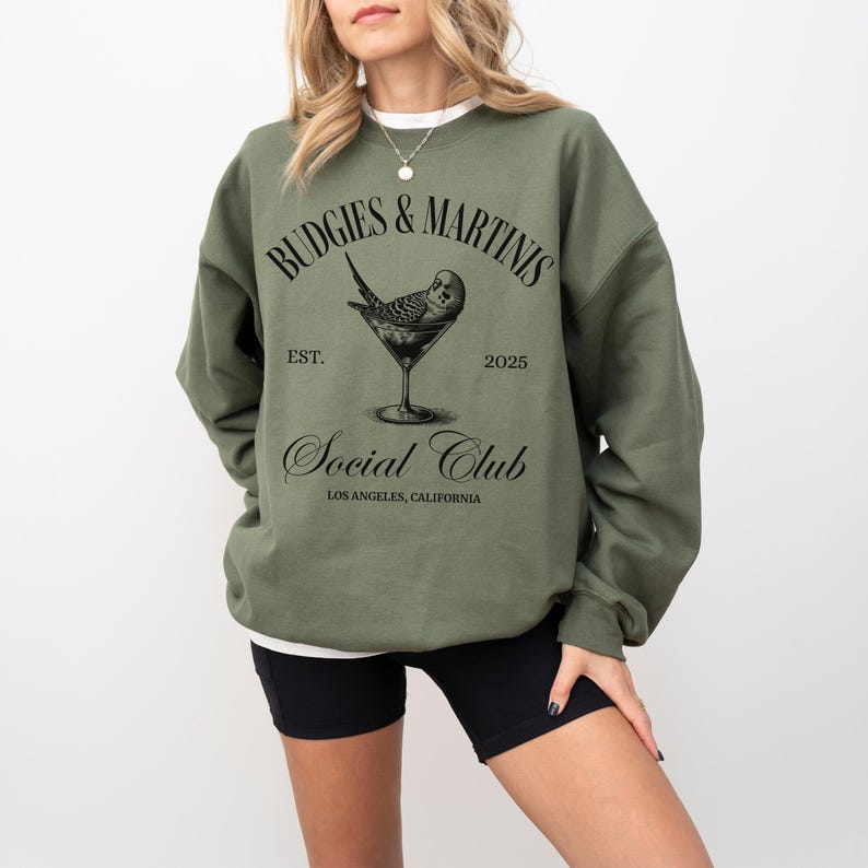May include: A green sweatshirt with the text "Budgies & Martins Social Club Est. 2025 Los Angeles, California" and a black and white illustration of a budgie in a martini glass.