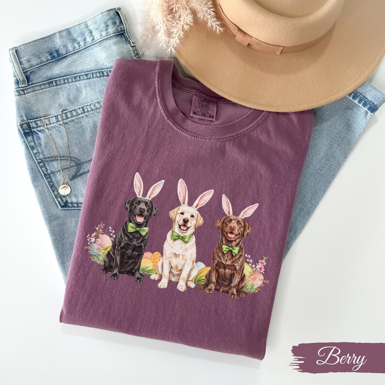 May include: A purple t-shirt with a graphic of three Labrador Retrievers wearing bunny ears and bow ties. The dogs are surrounded by Easter eggs. The shirt is called "Berry".