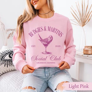 May include: A light pink sweatshirt with the text "Budgies & Martinis Social Club" in purple. The text is surrounded by a circle with a budgie perched on the rim of a martini glass.