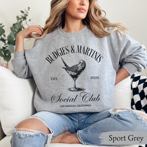 May include: A gray sweatshirt with the text "Budgies & Martinis Social Club Est. 2025 Los Angeles, California" and a black and white illustration of a budgie in a martini glass.