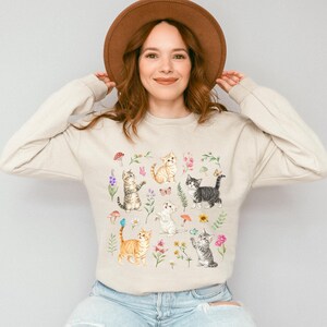 May include: A beige sweatshirt with a colorful print of cats and flowers. The cats are in various poses, including sitting, standing, and playing. The flowers are in bloom and have different colors, including pink, purple, and yellow.
