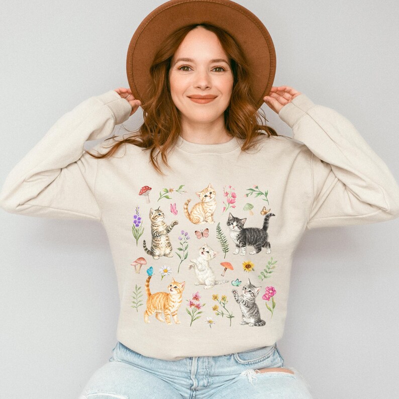 May include: A beige sweatshirt with a colorful print of cats and flowers. The cats are in various poses, including sitting, standing, and playing. The flowers are in bloom and have different colors, including pink, purple, and yellow.