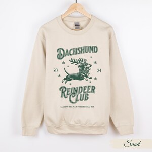 May include: A beige sweatshirt featuring a green graphic of a dachshund dressed as a reindeer. The graphic reads "Dachshund Reindeer Club" and "Leading the way to Christmas joy".