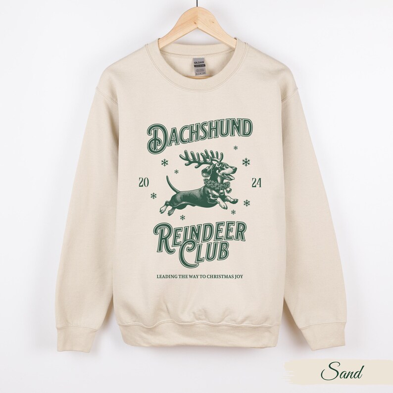 May include: A beige sweatshirt featuring a green graphic of a dachshund dressed as a reindeer. The graphic reads "Dachshund Reindeer Club" and "Leading the way to Christmas joy".
