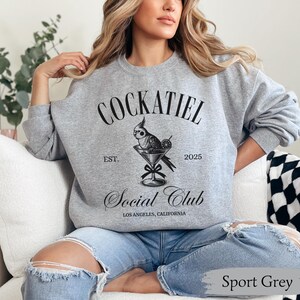 May include: A grey sweatshirt with the text "Cockatiel Social Club" in black. The design features a cockatiel perched on a martini glass with a lime wedge. The text "Est. 2025" is below the bird and the text "Los Angeles, California" is below the club name.