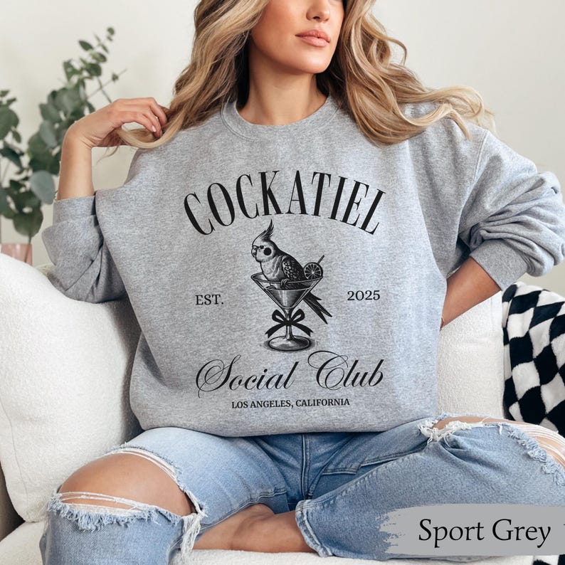 May include: A grey sweatshirt with the text "Cockatiel Social Club" in black. The design features a cockatiel perched on a martini glass with a lime wedge. The text "Est. 2025" is below the bird and the text "Los Angeles, California" is below the club name.