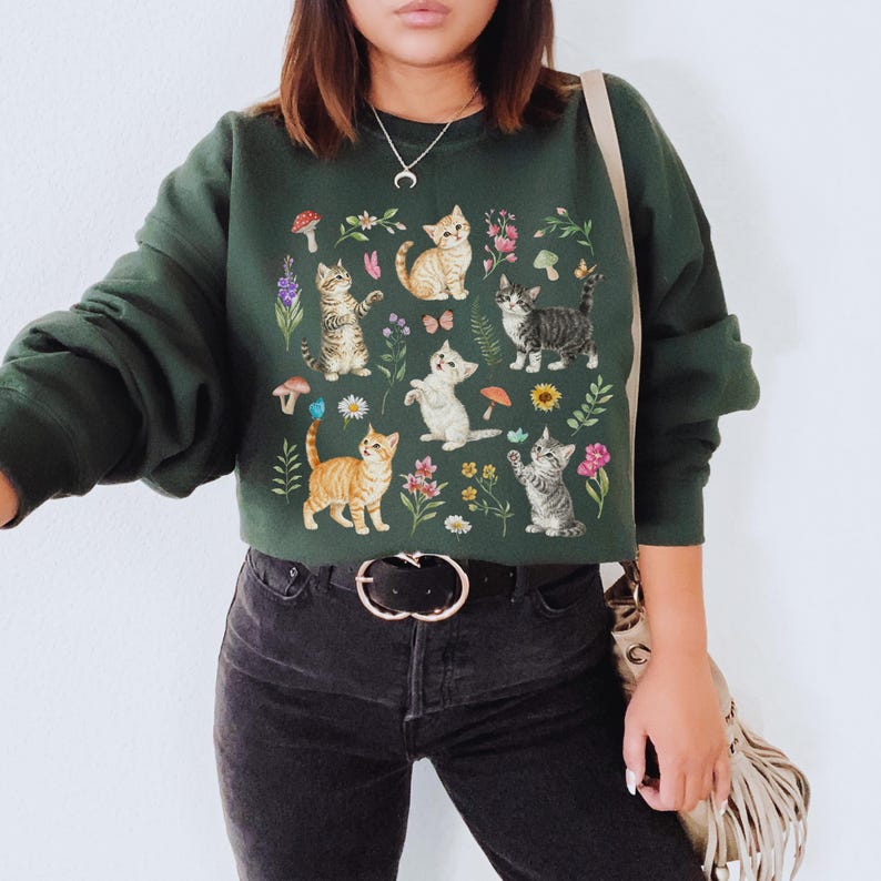 May include: A dark green sweatshirt with a print of cats and flowers. The cats are in various poses and colors, including white, orange, and gray. The flowers are in shades of pink, purple, and yellow.