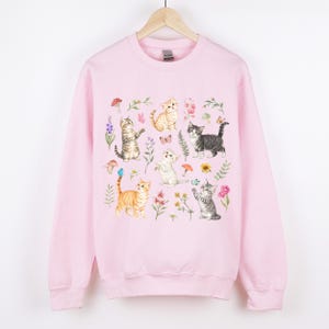 May include: Pink sweatshirt with a graphic print of multiple cats and flowers. The cats are in various poses and colors, including orange, gray, and white. The flowers are in shades of pink, purple, and yellow.