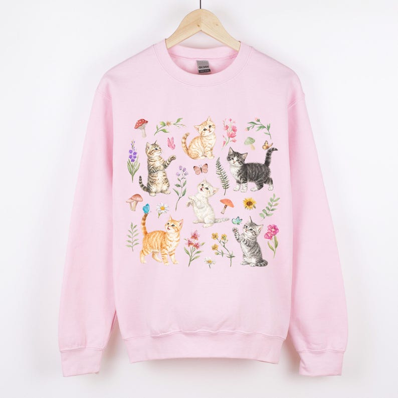 May include: Pink sweatshirt with a graphic print of multiple cats and flowers. The cats are in various poses and colors, including orange, gray, and white. The flowers are in shades of pink, purple, and yellow.