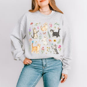 May include: A gray sweatshirt with a colorful print of cats and flowers. The cats are in various poses, some are sitting, some are standing, and some are playing. The flowers are in a variety of colors, including pink, purple, yellow, and blue.