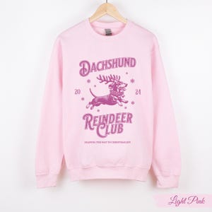 May include: Light pink sweatshirt featuring a pink dachshund wearing reindeer antlers and a festive collar. The text "Dachshund Reindeer Club" is printed on the sweatshirt, along with the words "Leading the way to Christmas joy".