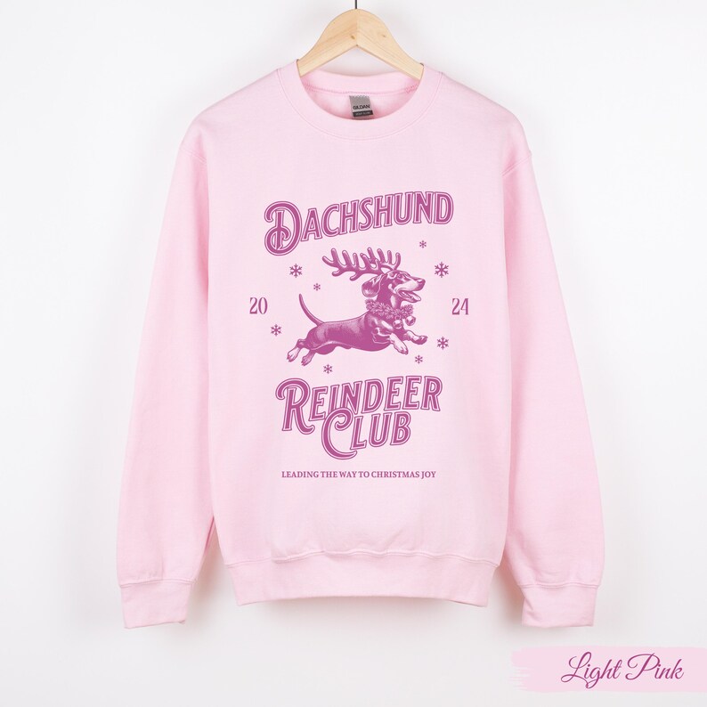 May include: Light pink sweatshirt featuring a pink dachshund wearing reindeer antlers and a festive collar. The text "Dachshund Reindeer Club" is printed on the sweatshirt, along with the words "Leading the way to Christmas joy".