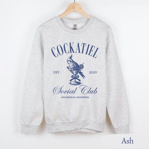 May include: A grey sweatshirt featuring a blue graphic of a cockatiel in a martini glass. The text "Cockatiel Social Club" is printed on the sweatshirt, along with "Est. 2025" and "Los Angeles, California". The sweatshirt is labelled "Ash".