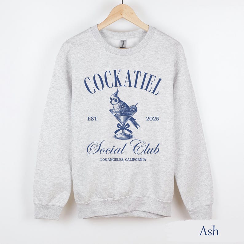 May include: A grey sweatshirt featuring a blue graphic of a cockatiel in a martini glass. The text "Cockatiel Social Club" is printed on the sweatshirt, along with "Est. 2025" and "Los Angeles, California". The sweatshirt is labelled "Ash".