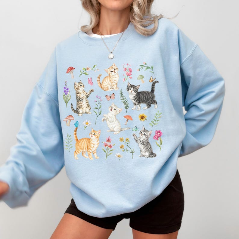 May include: Light blue sweatshirt with a colorful print of kittens playing in a field of flowers. The kittens are in various poses, including sitting, standing, and jumping. The flowers are in shades of pink, purple, and yellow.