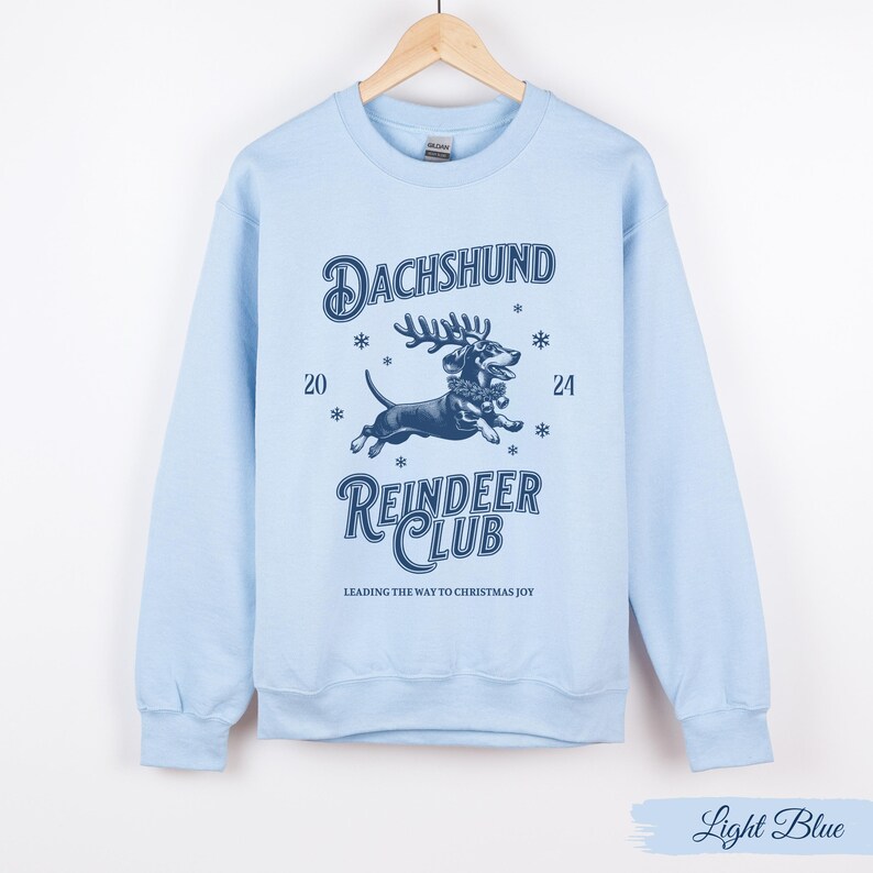 May include: Light blue sweatshirt featuring a graphic of a dachshund wearing reindeer antlers and the text "Dachshund Reindeer Club" and "Leading the way to Christmas joy".