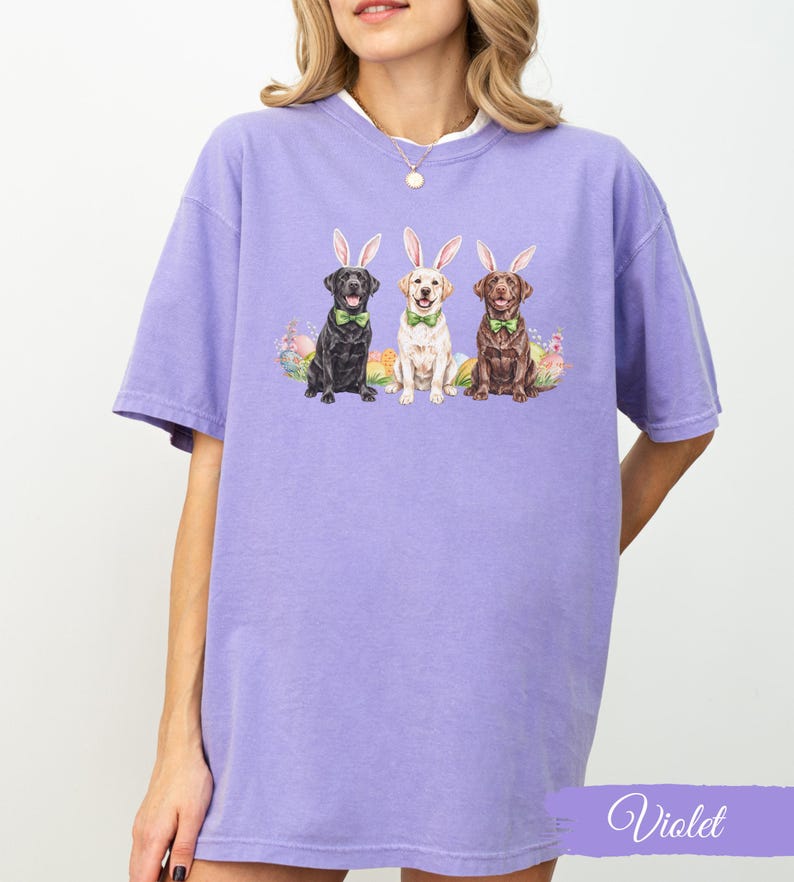 May include: A purple t-shirt with a graphic of three Labrador Retrievers wearing bunny ears and bow ties. The dogs are sitting in front of a background of Easter eggs.