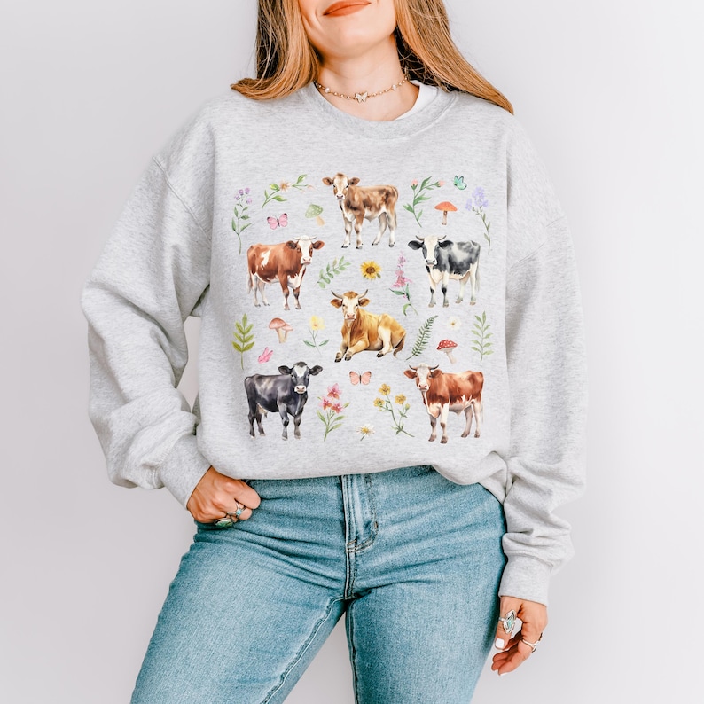 Pu&ograve; includere: Una felpa grigia con una stampa colorata di mucche, fiori e funghi. Le mucche sono in varie pose e colori, tra cui marrone, nero e bianco.