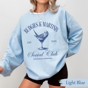 May include: Light blue sweatshirt with the text "Budgies & Martinis Social Club Est. 2025 Los Angeles, California" and a bluebird in a martini glass.