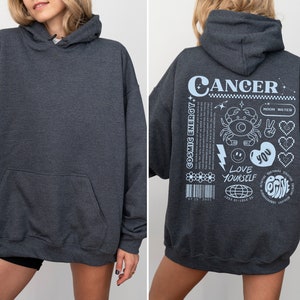 May include: A dark gray hooded sweatshirt with a white graphic on the back that says "Cancer" and includes a crab, stars, hearts, and the text "Cosmic Energy", "Love Yourself", and "You".