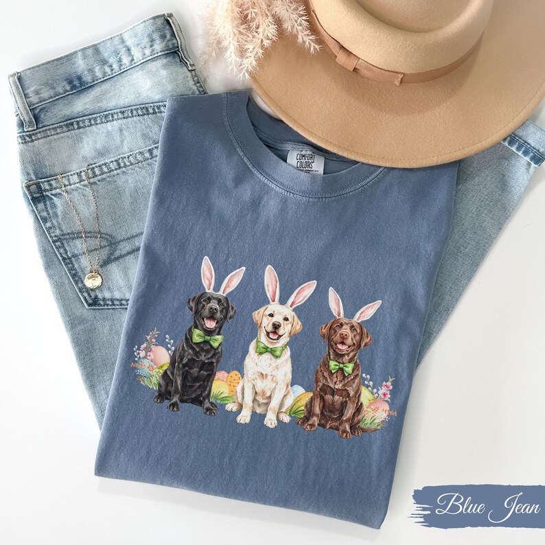 May include: A blue jean t-shirt with three Labrador Retrievers wearing bunny ears. The dogs are wearing green bow ties and are surrounded by Easter eggs. The text "Comfort Colors" is visible on the shirt.