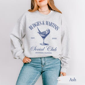 May include: A gray sweatshirt with the text "Budgies & Martins Social Club Est. 2025 Los Angeles, California" and a blue illustration of a budgie in a martini glass.