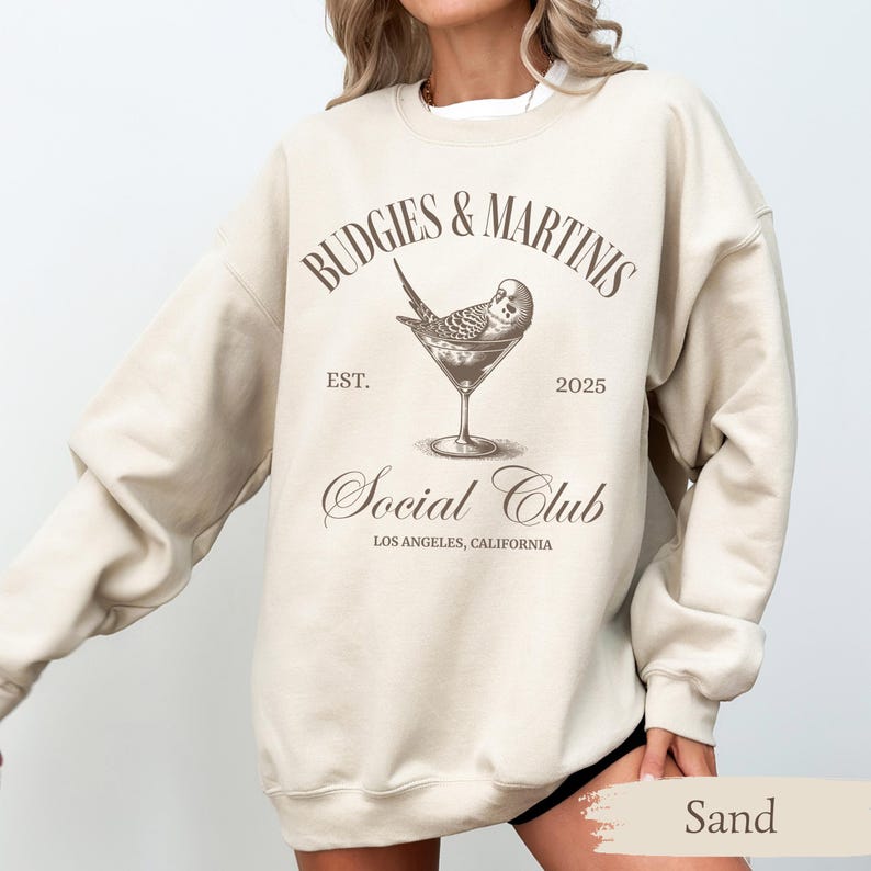 May include: A beige sweatshirt with the text "Budgies & Martinis Social Club Est. 2025 Los Angeles, California" and a graphic of a budgie in a martini glass.