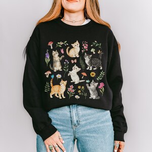 May include: Black sweatshirt with a colorful print of kittens and flowers. The kittens are in various poses, including sitting, standing, and playing. The flowers are in shades of pink, purple, and yellow.