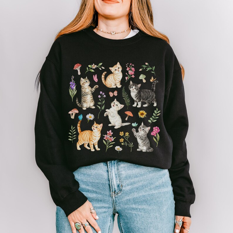 May include: Black sweatshirt with a colorful print of kittens and flowers. The kittens are in various poses, including sitting, standing, and playing. The flowers are in shades of pink, purple, and yellow.