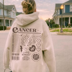 May include: A beige hoodie with a graphic design featuring the zodiac sign Cancer, the text "Cancer", and other astrological symbols. The design also includes the text "Cosmic Energy", "Moon Water", "Love Yourself", and "Positive".