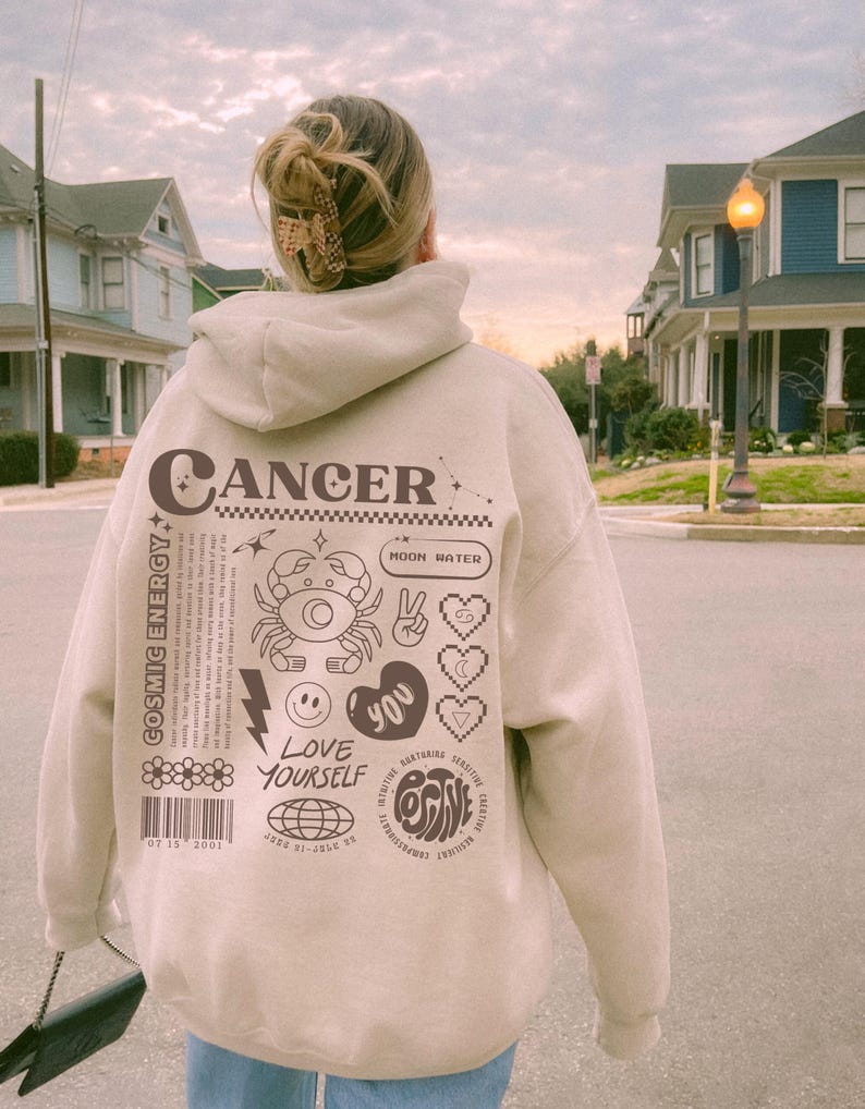 May include: A beige hoodie with a graphic design featuring the zodiac sign Cancer, the text "Cancer", and other astrological symbols. The design also includes the text "Cosmic Energy", "Moon Water", "Love Yourself", and "Positive".