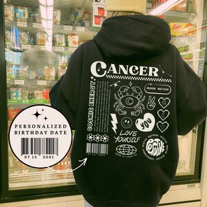 May include: Black hoodie with a white graphic print of the zodiac sign Cancer. The graphic includes the words "Cosmic Energy", "Moon Water", "Love Yourself", and "Positive". The hoodie also has a personalized birthday date barcode.
