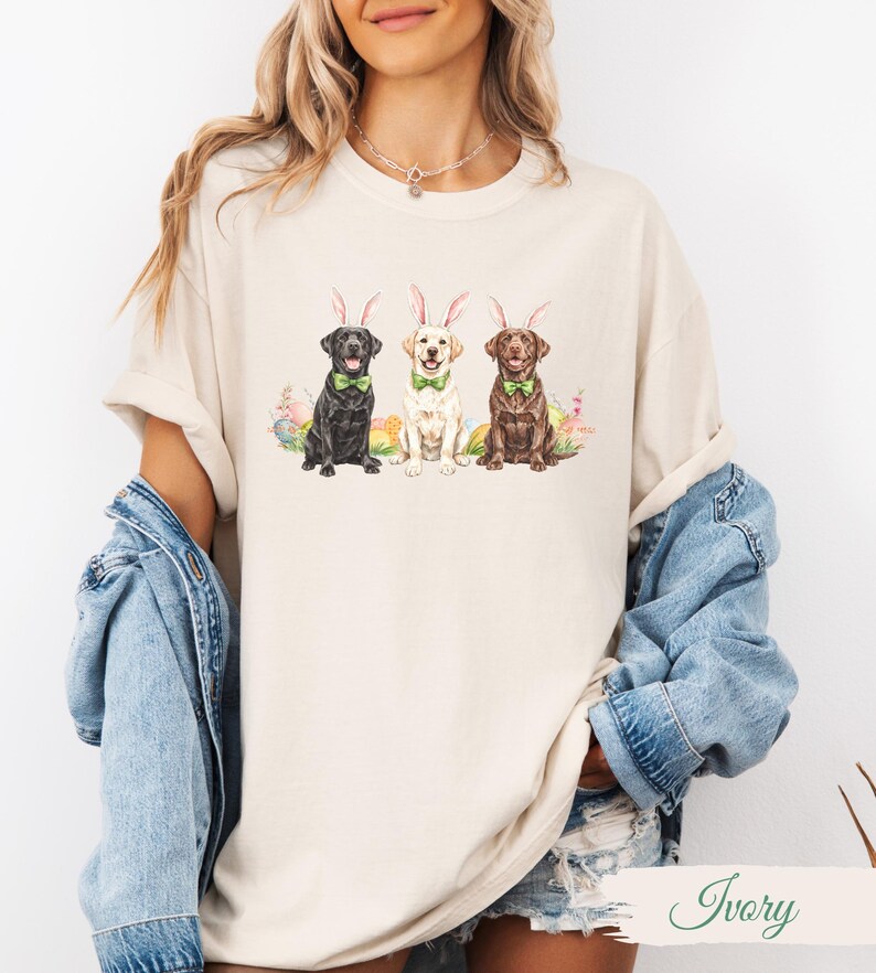 May include: A white t-shirt with a graphic of three Labrador Retrievers wearing bunny ears. The dogs are black, white, and brown. The graphic is surrounded by pastel colored Easter eggs.