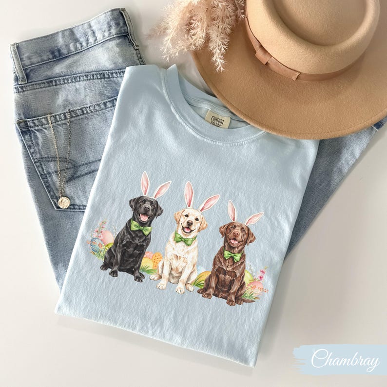 May include: A light blue t-shirt with a graphic of three Labrador Retrievers wearing bunny ears. The dogs are surrounded by Easter eggs and flowers. The text "Chambray" is visible in the bottom right corner of the image.