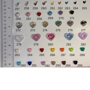 May include: A collection of heart-shaped charms and beads arranged in rows, labelled with numbers 253 through 295. A clear plastic ruler is positioned on the left side, showing measurements in inches up to 8 inches for scale reference.